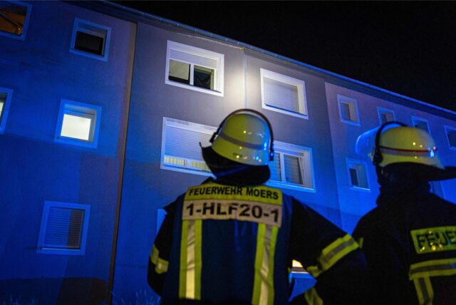 Zwei Personen der Feuerwehr stehen vor einem Mehrfamilienhaus und schauen hoch zu einem Fenster.