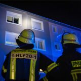 Zwei Personen der Feuerwehr stehen vor einem Mehrfamilienhaus und schauen hoch zu einem Fenster.