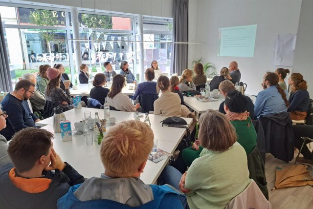 Im Hertener Projektbüro Neustart Innenstadt an der Ewaldstraße sitzen Männer und Frauen an mehreren Tischen und blicken auf eine Projektion an der Wand.