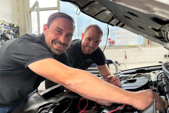 Simon Dzierzon und Jochen Anufriev stehen an der geöffneten Motorhaube eines Autos. Die Kamera steht neunzig Grad zur Motorhaube, am Kotflügel. Dzierzon steht neben Anufriev und schließt ein Lesegerät an die Schnittstelle im Motor an. Beide lächeln in die Kamera.
