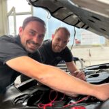 Simon Dzierzon und Jochen Anufriev stehen an der geöffneten Motorhaube eines Autos. Die Kamera steht neunzig Grad zur Motorhaube, am Kotflügel. Dzierzon steht neben Anufriev und schließt ein Lesegerät an die Schnittstelle im Motor an. Beide lächeln in die Kamera.