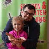 Ein Mann hat ein Mädchen auf dem Schoß. Beide halten eine Vogelspinne in den Händen und lächeln in die Kamera. Im Hintergrund ist ein Terrarium zu erkennen.