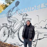 Norbert Smuda steht vor einem der Motive des neuen Graffiti an der Allee des Wandels. Es zeigt eine Person auf einem Fahrrad, die eine Straße entlang schaut. Das Motiv ist in schwarz auf einem hellbeigen Grund gesprayed und wird von einem hellblauen Schriftzug „Allee des Wandels“ durchbrochen. Die Schultern der Personen sind ebenfalls vor einem hellblauen Hintergrund. Am oberen linken Rand des Bildes sind herbstliche Bäume über der Mauer zu erkennen.
