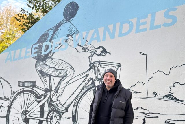 Norbert Smuda steht vor einem der Motive des neuen Graffiti an der Allee des Wandels. Es zeigt eine Person auf einem Fahrrad, die eine Straße entlang schaut. Das Motiv ist in schwarz auf einem hellbeigen Grund gesprayed und wird von einem hellblauen Schriftzug „Allee des Wandels“ durchbrochen. Die Schultern der Personen sind ebenfalls vor einem hellblauen Hintergrund. Am oberen linken Rand des Bildes sind herbstliche Bäume über der Mauer zu erkennen.