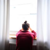 Eine Frau sitzt mit Kopf in den Händen am Fenster.