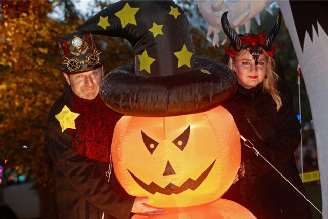 Zwei kostümierte Menschen stehen an Halloween neben einem beleuchteten Kürbis, der einen Hexenhut trägt.