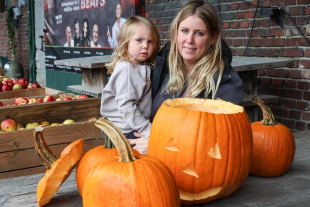 Kristina Mertin, eine junge Frau, sitzt gemeinsam mit ihrem Kind vor einem Tisch, auf dem Kürbisse liegen. Einer von den Halloweenkürbissen, hat ein geschnitztes Gesicht.