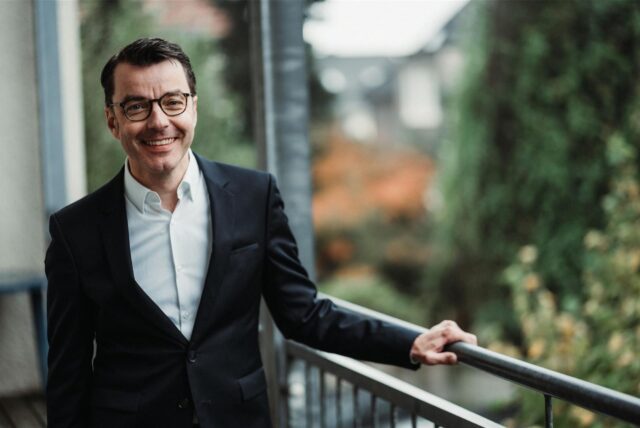 SPD-Landratskandidat Karsten Schneider trägt Sakko und Hemd. Er lehnt an einem Geländer und lächelt in die Kamera.