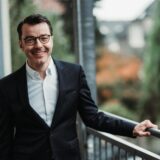 SPD-Landratskandidat Karsten Schneider trägt Sakko und Hemd. Er lehnt an einem Geländer und lächelt in die Kamera.