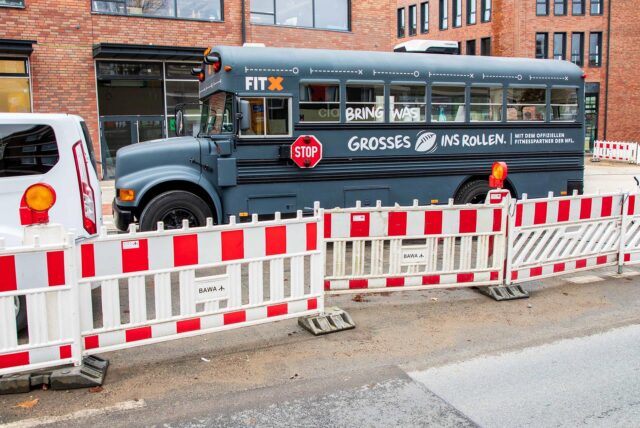 Hinter einer rot-weißen Baustellen-Absperrung steht ein grauer Bus vor den Hertener Höfen. An der seitlichen Karosserie steht in weißer Schrift „Bring was Großes ins Rollen“. Zwischen „Großes“ und „ins“ ist ein Football abgebildet. ETwas weiter vorne an der Seite hängt ein Stop-Schild. Über dem Fahrerfenster steht „FitX“.