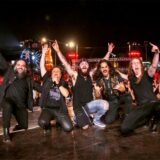 Fünf Musiker nebeneinander auf der Bühne, die für die Kamera posieren und die Arme hochreißen, hinter ihnen jubelnde Fans.