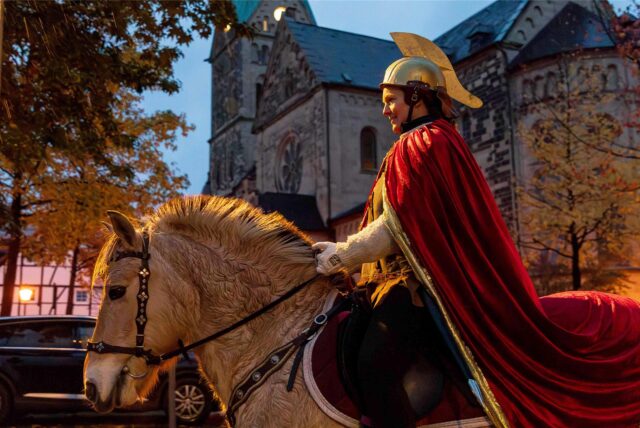 Katrin Zobel reitet als Sankt Martina auf einem Pferd durch das Alte Dorf Westerholt in Herten. Im Hintergrund ist die St.Martinus-Kirche zu sehen.