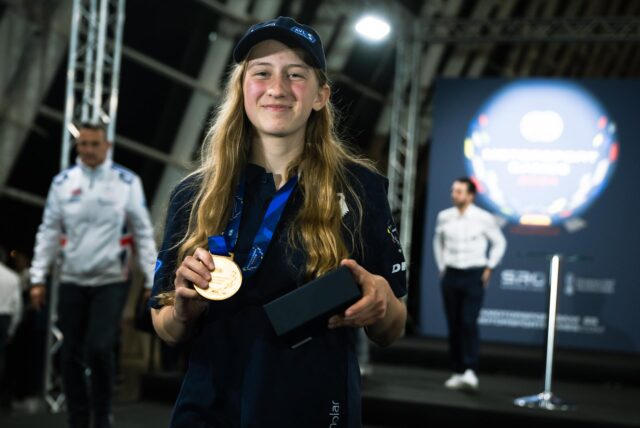 Maja Braun steht mit ihrer Goldmedaille in der Veranstaltungshalle in Valencia. Im Hintergrund ist das Logo der FIA Motorsport Games zu erkennen. Mehrere Personen laufen durch die Halle. Maja trägt eine blaue Kappe und ein dunkles Poloshirt.
