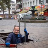 Thorsten Raschke ragt halb aus einem Abwasserkanal hervor, im Hintergrund ist der Brunnen an der Kranzplatte zu sehen.