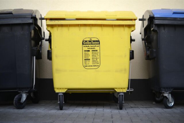 Ein gelber Müllcontainer steht vor einer beigen Hauswand. Das „Gelbe Tonne“ Logo ist deutlich auf der Vorderseite zu erkennen. Links und rechts steht eine schwarze Restmülltonnen und eine Papiertonne, zu erkennen am blauen Deckel.