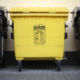 Ein gelber Müllcontainer steht vor einer beigen Hauswand. Das „Gelbe Tonne“ Logo ist deutlich auf der Vorderseite zu erkennen. Links und rechts steht eine schwarze Restmülltonnen und eine Papiertonne, zu erkennen am blauen Deckel.