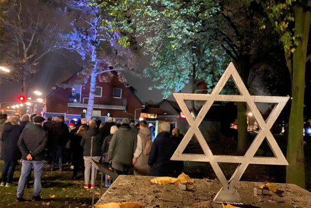 Menschen hören einem Redner zu. Im Vordergrund steht ein Judenstern aus Holz.