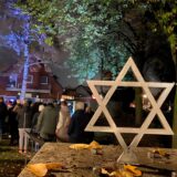 Menschen hören einem Redner zu. Im Vordergrund steht ein Judenstern aus Holz.
