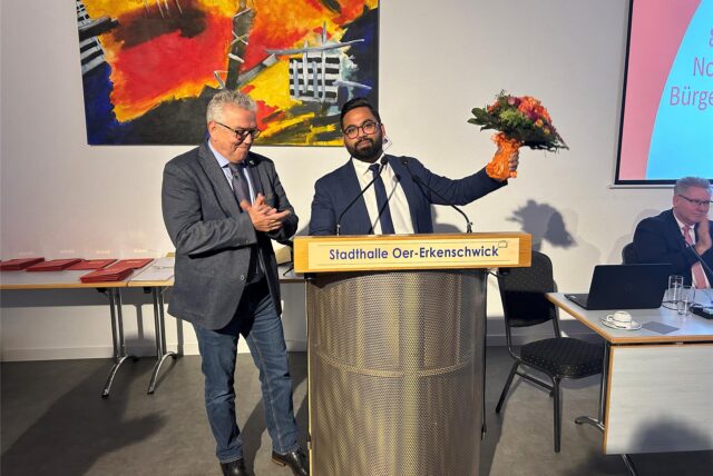 Ein Mann in Anzug und Krawatte hält einen Blumenstrauß in der linken Hand. Ein Mann neben ihm klatscht in die Hände.