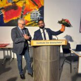 Ein Mann in Anzug und Krawatte hält einen Blumenstrauß in der linken Hand. Ein Mann neben ihm klatscht in die Hände.