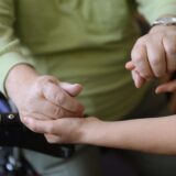 Eine Person in einem Grünen Pullover wird von einer anderen Person an die Hände genommen. Man sieht nur den Torso der einen Person und die zwei Arme der anderen. Die Person im Grünen Pullover stützt ihre Hand auf den Griff eines Rollators.