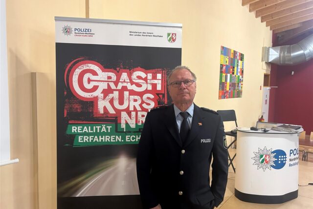 Martin Weinhold steht in schwarzer Polizeiuniform auf der Bühne in der Mensa des Willy-Brandt-Gymnasiums in Oer-Erkenschwick. Im Hintergrund ist ein Plakataufsteller mit der Aufschrift „Crash Kurs NRW“.