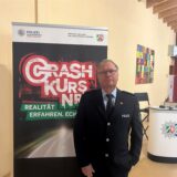 Martin Weinhold steht in schwarzer Polizeiuniform auf der Bühne in der Mensa des Willy-Brandt-Gymnasiums in Oer-Erkenschwick. Im Hintergrund ist ein Plakataufsteller mit der Aufschrift „Crash Kurs NRW“.