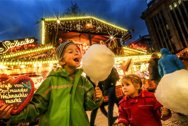 Ein Junge steht neben einem kleinen Mädchen auf dem Dortmunder Weihnachtsmarkt. Beide halten eine große Zuckerwatte in der Hand, der Junge hält außerdem ein Lebkuchenherz in die Kamera.