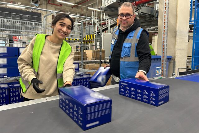 Christoph Brings, Standortleiter bei Arvato in Dorsten, steht neben einer Mitarbeiterin an einem Fließband, auf das die Bestellungen gelegt werden.