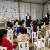 Regierungspräsident Andreas Bothe sitzt vor vielen Kindern, die auf weißen Stühlen sitzen.