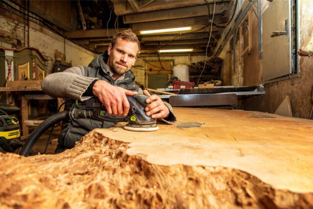 Florian Karwig kniet in seiner Werkstatt auf dem Spargelhof Schulte-Scherlebeck in Herten an einer großen Holzplatte und hält eine Schleifmaschine in der Hand.