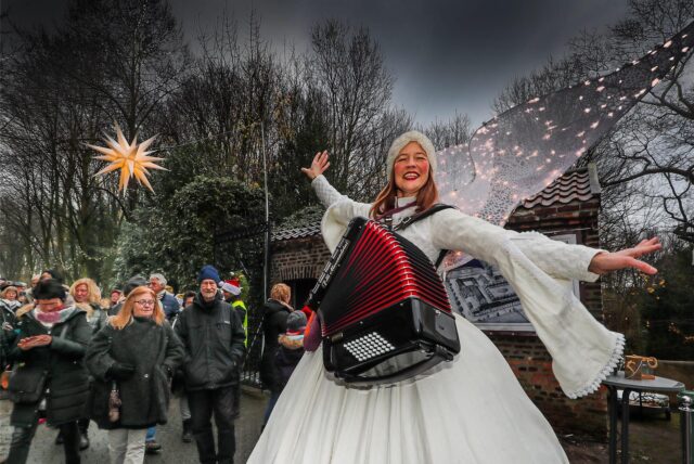 Eine Frau, als Engel verkleidet und mit Ziehharmonika um den Hals, lächelt in die Kamera. Sie steht auf dem Marktplatz des Weihnachtsmarkts auf dem Schloss Bodelschwingh in Dortmund.