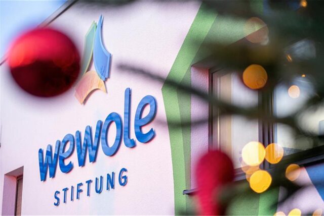 Das Logo der Wewole-Stiftung ist abgebildet. Leicht verschwommen sind davor die Lichter einer Lichterkette und eine rote Weihnachtskugel zu erkennen.
