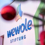 Das Logo der Wewole-Stiftung ist abgebildet. Leicht verschwommen sind davor die Lichter einer Lichterkette und eine rote Weihnachtskugel zu erkennen.