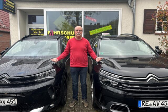 Ein Mann in rotem Pullover steht neben zwei schwarzen Autos vor seiner Fahrschule in Oer-Erkenschwick.