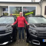 Ein Mann in rotem Pullover steht neben zwei schwarzen Autos vor seiner Fahrschule in Oer-Erkenschwick.