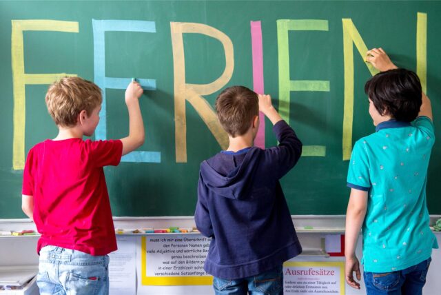 Drei Kinder schreiben das Wort Ferien mit bunter Kreide an die Tafel.