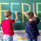 Drei Kinder schreiben das Wort Ferien mit bunter Kreide an die Tafel.