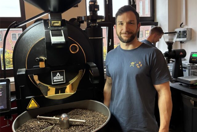 Dominik Schulte Sasse steht rechts neben einer Kaffeeröstmaschine vom Hersteller Probat. In der Kühltrommel liegen Kaffeebohnen. Hinter ihm steht ein Mitarbeiter an einer Kaffeemühle.