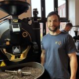 Dominik Schulte Sasse steht rechts neben einer Kaffeeröstmaschine vom Hersteller Probat. In der Kühltrommel liegen Kaffeebohnen. Hinter ihm steht ein Mitarbeiter an einer Kaffeemühle.