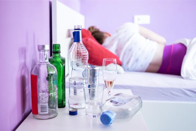 Eine Frau liegt im Bett – vor ihr mehrere leere Flaschen auf einem Nachttisch.