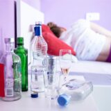Eine Frau liegt im Bett – vor ihr mehrere leere Flaschen auf einem Nachttisch.