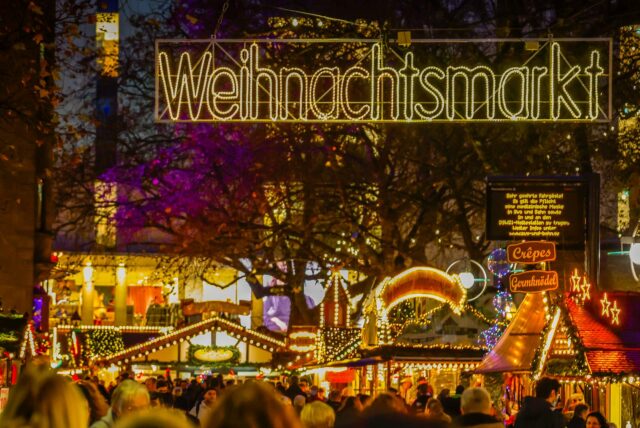 Ein Leuchtschild mit dem Wort „Weihnachtsmarkt.“ Menschen schlendern zwischen den Buden entlang.