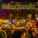 Ein Leuchtschild mit dem Wort „Weihnachtsmarkt.“ Menschen schlendern zwischen den Buden entlang.