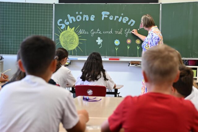 Eine Lehrerin schreibt „Schöne Ferien“ mit Kreide an eine Schultafel und die Schüler sitzen an den Tischen.