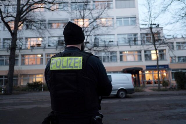 Ein Polizist hat der Kamera den Rücken zugedreht und steht vor einem großen Gebäude.