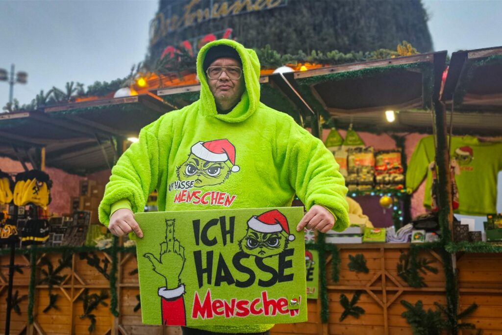 Christoph Scheiding steht vor seinem Stand auf dem Dortmunder Weihnachtsmarkt und zeigt eine Fußmatte mit der Aufschrift Christoph Scheiding steht vor seinem Stand auf dem Dortmunder Weihnachtsmarkt und zeigt eine Fußmatte mit der Aufschrift