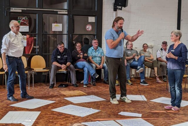 Menschen in der Maschinenhalle, eine Person hält ein Mikrofon in der Hand und gestikuliert.
