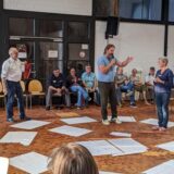 Menschen in der Maschinenhalle, eine Person hält ein Mikrofon in der Hand und gestikuliert.