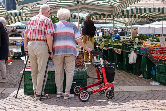 Zwei Senioren stehen mit Gehhilfen und Rollator auf dem Wochenmarkt.
